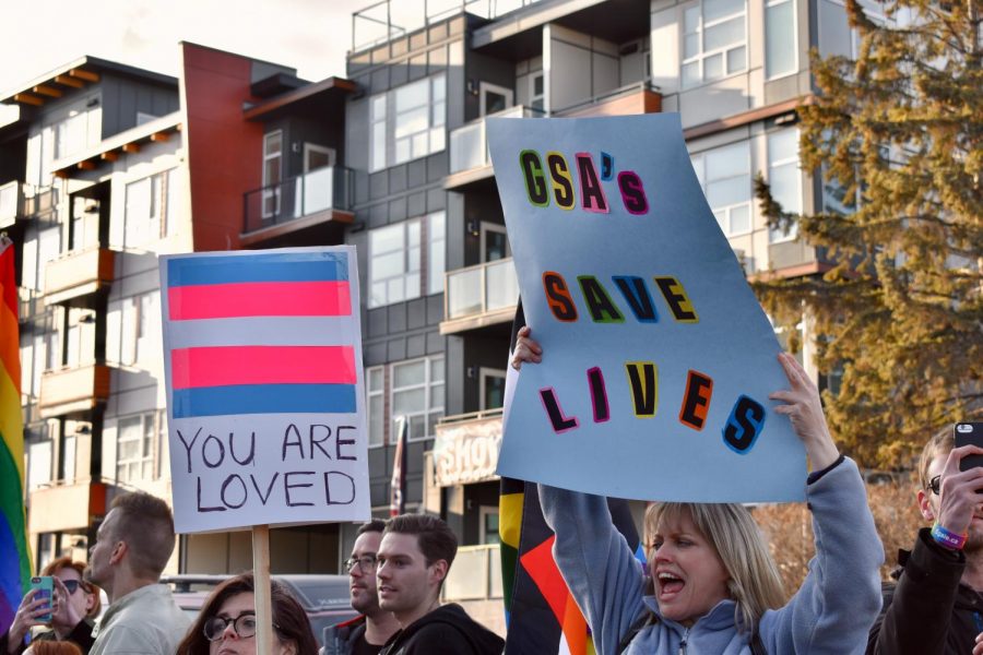 A group of protesters holds signs reading GSAs Save Lives and Your Are Loved beneath a trans flag.